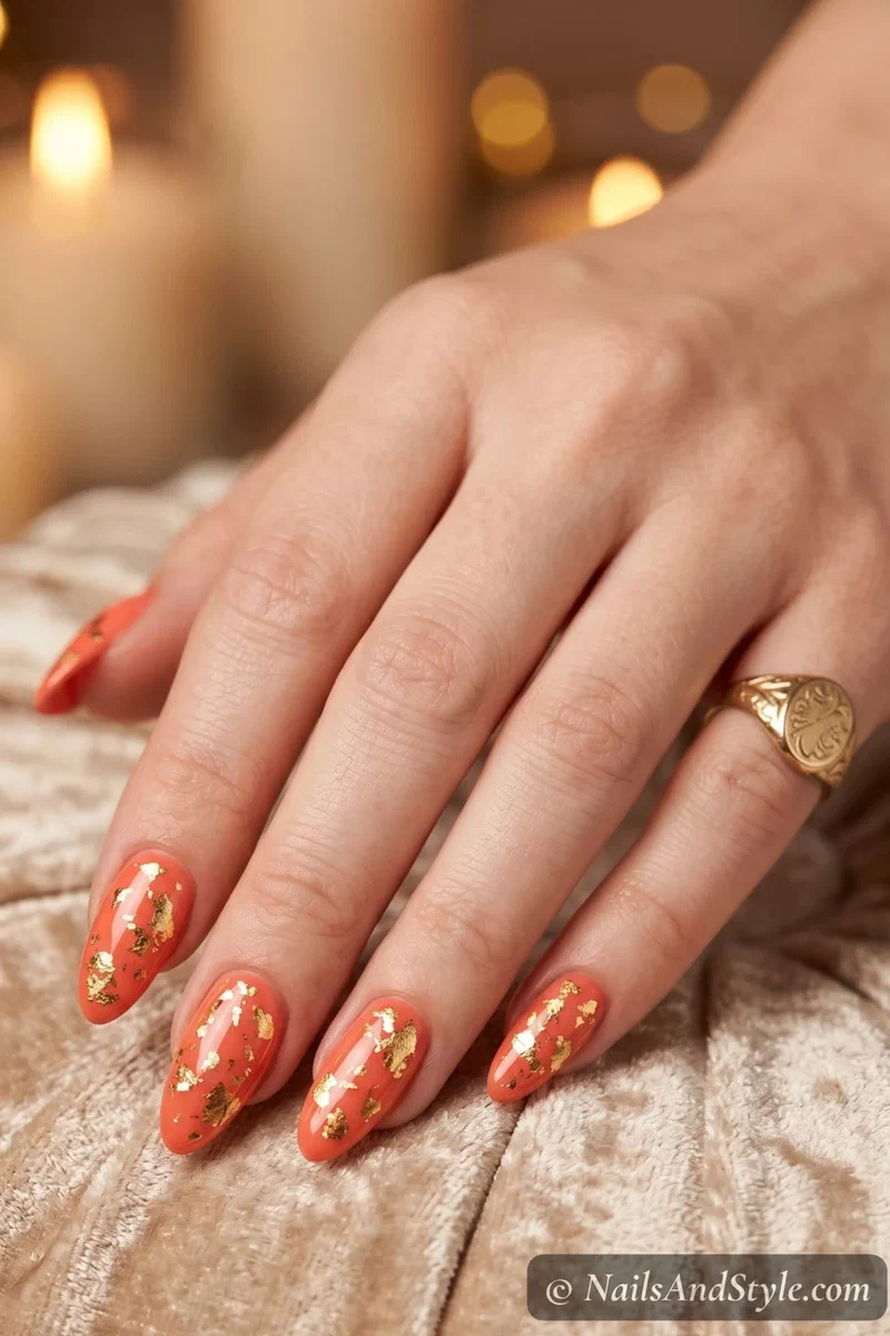 Warm coral nails with scattered gold foil accents on medium almond nails